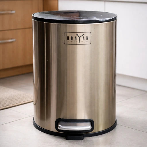 Stainless steel trash can with black lid and pedal on a kitchen floor.