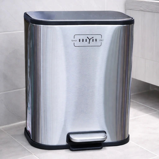 Stainless steel trash bin with black lid and foot pedal on a tiled floor.