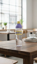 Clear bottle with a purple label on a wooden table in a room with large windows.