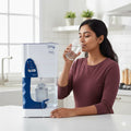 Woman drinking water from a glass next to a water dispenser in a kitchen.