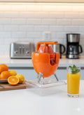 Orange manual juicer on a kitchen counter with oranges and a glass of juice.