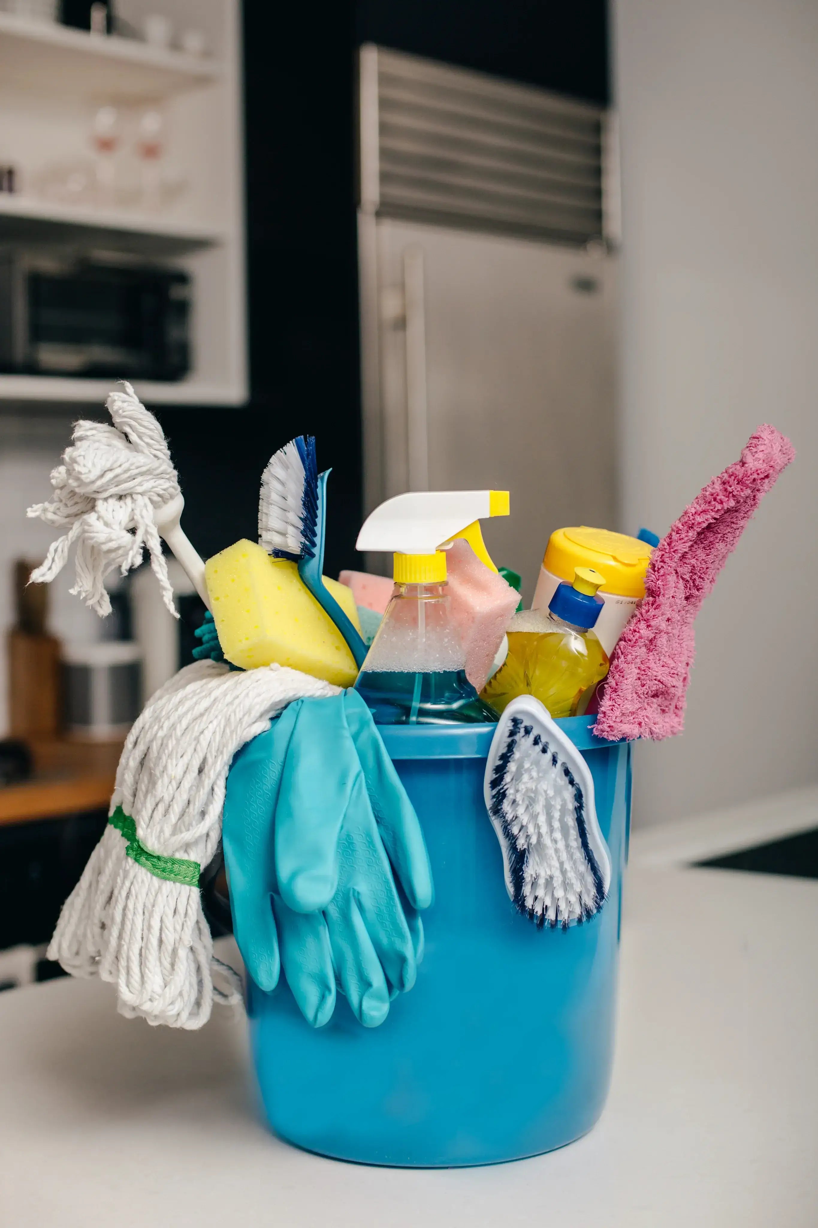 Blue cleaning bucket filled with mop, gloves, scrub brushes, sponges, and cleaning spray bottles placed on a kitchen countertop